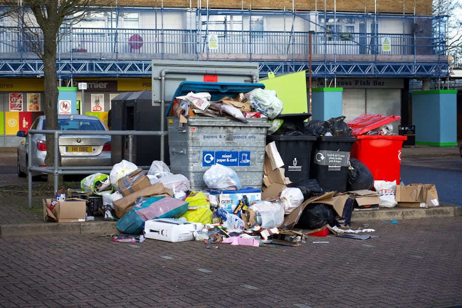 A collection of black bin bags, some ripped open, piled up against a textured, off-white wall near a red metal door covered with graffiti and various stickers, including a 'Keep Clear' sign and one in