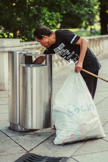 A large green wheeled waste bin filled with black and red bags containing construction or garden materials, positioned on a paved surface against a dark brick wall. The bags are marked with red and wh